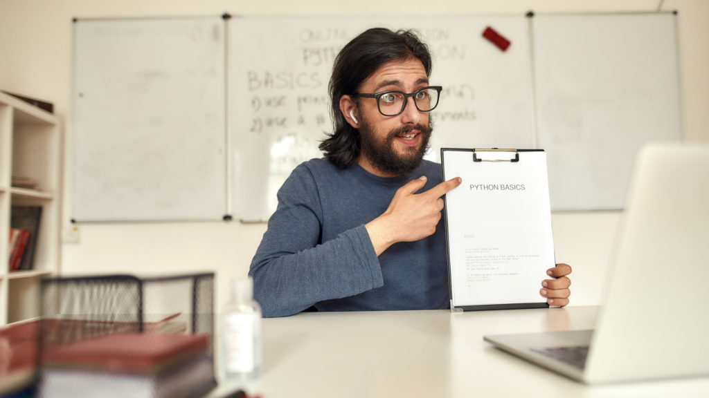Young bearded male teacher of code on his laptop with whiteboard in background.