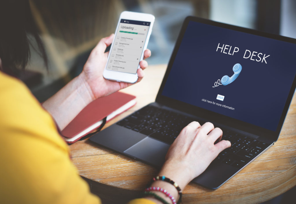 A woman in a yellow sweater using her phone and laptop to contact a Help Desk Support Technician.