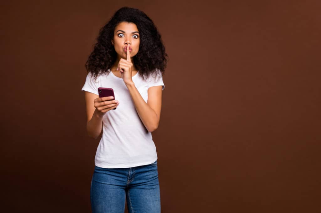Woman holding smartphone with finger on lips since she knows how to hide numbers on your phone bill