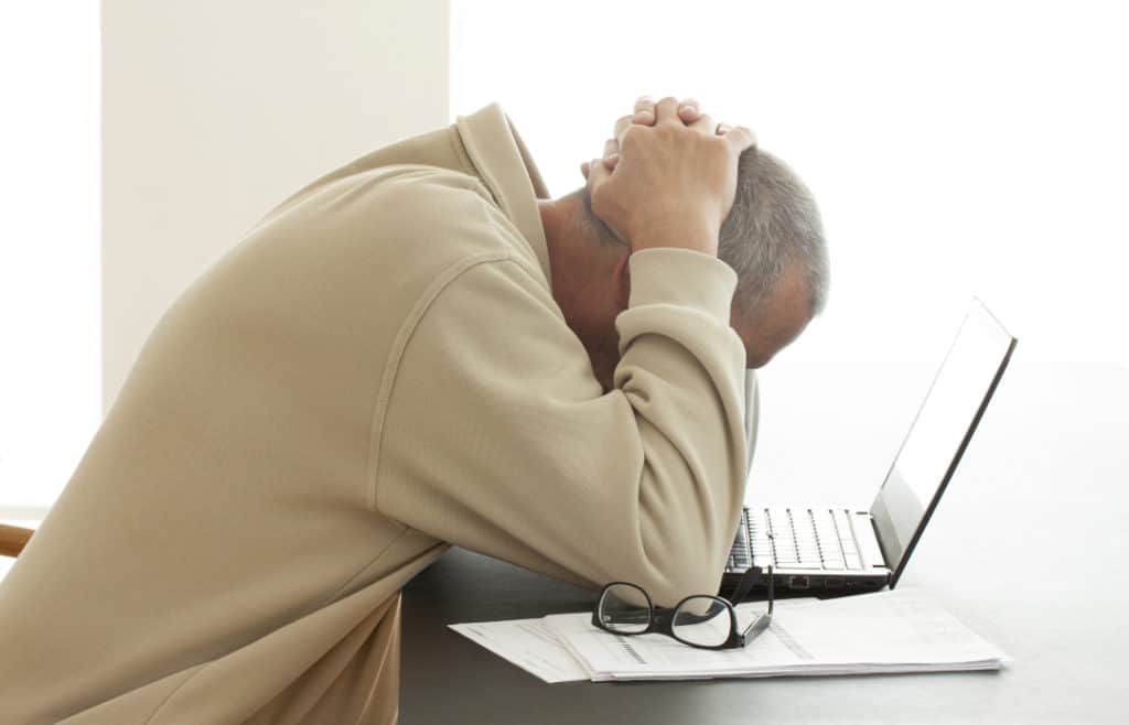 Man hands on head sadly looking down at laptop having clicked on attachment link after opening spam email