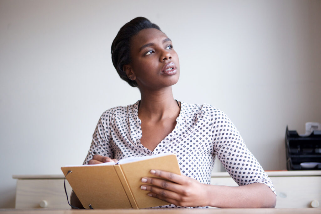 Serious young woman thinking and writing in a journal, considering options for using password generator words to create memorable passwords.